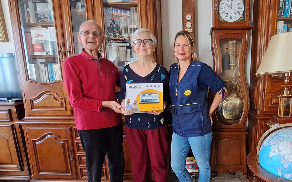 Deux personnes âgées recevant une tablette Ardoiz de La Poste, remise par une employée en uniforme, dans un salon avec des meubles en bois.