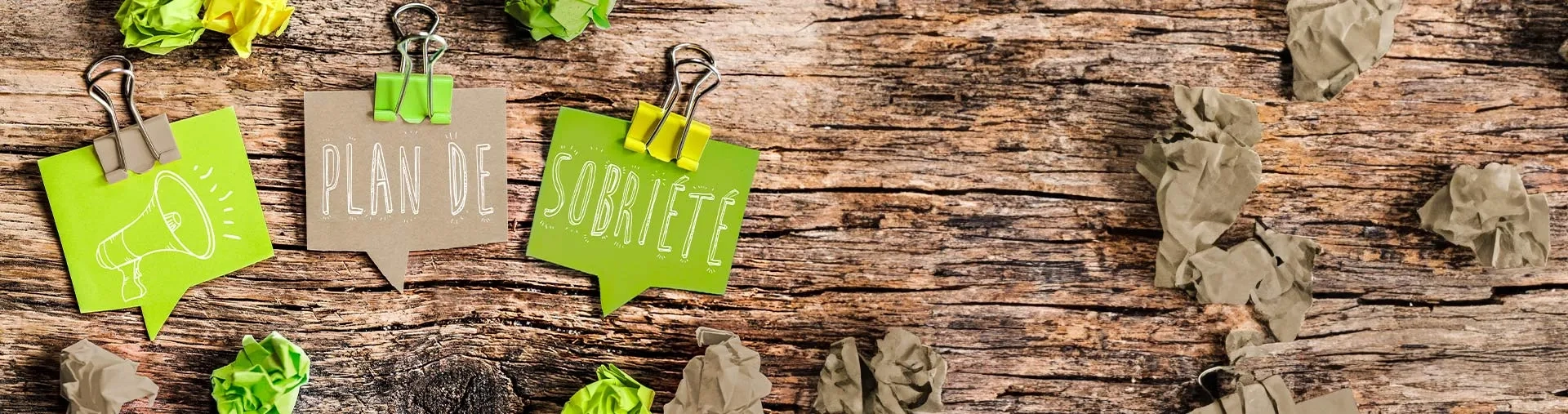 Sur un fond en bois texturé, il y a trois morceaux de papier avec des pinces de couleur verte. L'un montre une icône de mégaphone, tandis que les deux autres portent les mots "PLAN DE" et "SOBRIÉTÉ" écrits en lettres blanches.