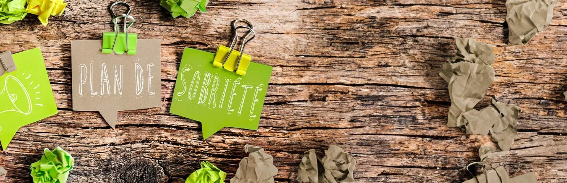 Sur un fond en bois texturé, il y a trois morceaux de papier avec des pinces de couleur verte. L'un montre une icône de mégaphone, tandis que les deux autres portent les mots "PLAN DE" et "SOBRIÉTÉ" écrits en lettres blanches.
