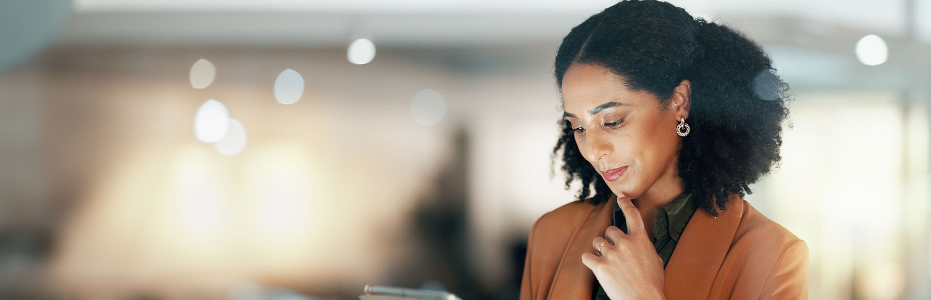  une femme professionnelle avec des cheveux bouclés, portant un blazer beige et des boucles d'oreilles dorées. Elle regarde attentivement une tablette, le fond flou avec des lumières bokeh crée une ambiance chaleureuse et moderne.