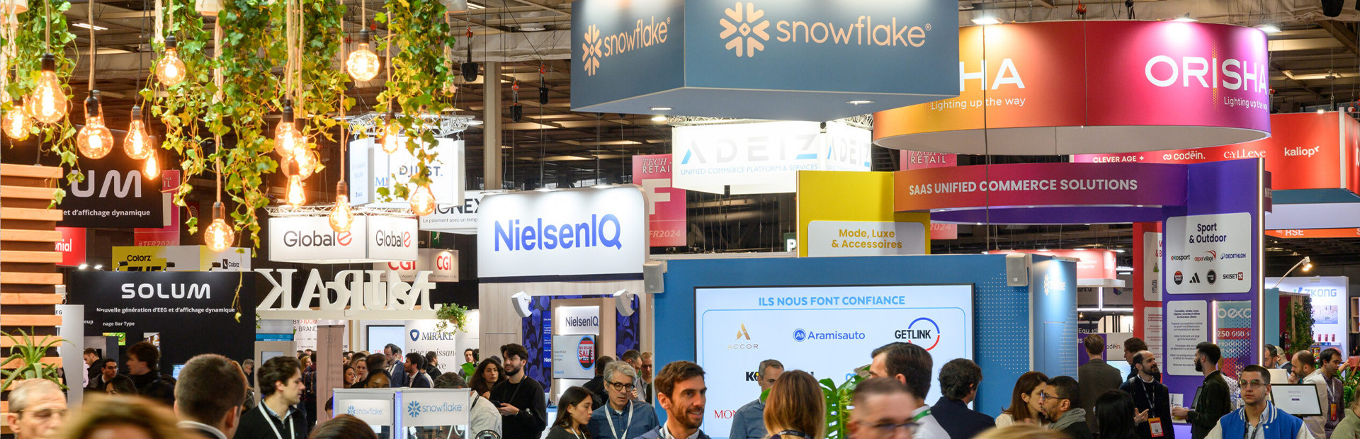 Vue d'un salon professionnel animé avec des stands d'entreprises, notamment Snowflake, NielsenIQ et Orisha. L'espace est décoré avec des plantes suspendues et éclairé par des ampoules chaleureuses. De nombreuses personnes discutent et se déplacent dans les allées