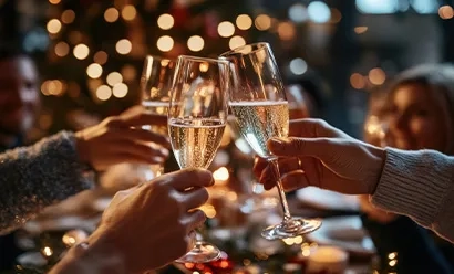 L'image montre deux mains tenant des coupes de champagne, s'entrechoquant dans un toast festif sur fond d'arbre de Noël et de lumières chaudes et scintillantes.