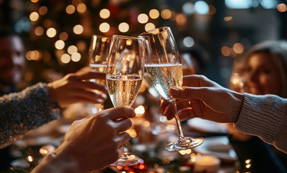 L'image montre deux mains tenant des coupes de champagne, s'entrechoquant dans un toast festif sur fond d'arbre de Noël et de lumières chaudes et scintillantes.