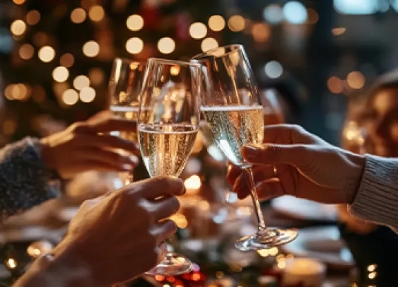 L'image montre deux mains tenant des coupes de champagne, s'entrechoquant dans un toast festif sur fond d'arbre de Noël et de lumières chaudes et scintillantes.