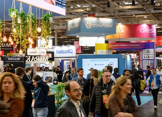 Vue d'un salon professionnel animé avec des stands d'entreprises, notamment Snowflake, NielsenIQ et Orisha. L'espace est décoré avec des plantes suspendues et éclairé par des ampoules chaleureuses. De nombreuses personnes discutent et se déplacent dans les allées