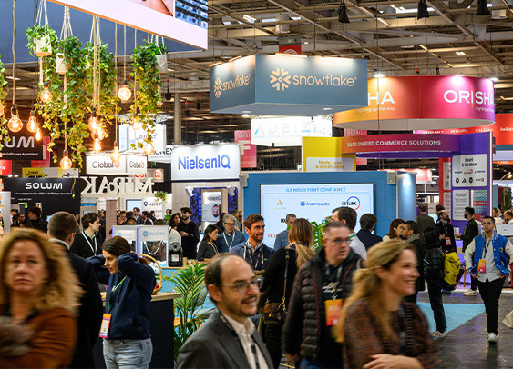 Vue d'un salon professionnel animé avec des stands d'entreprises, notamment Snowflake, NielsenIQ et Orisha. L'espace est décoré avec des plantes suspendues et éclairé par des ampoules chaleureuses. De nombreuses personnes discutent et se déplacent dans les allées